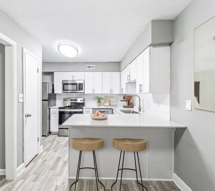 Modern kitchen with white shaker-style cabinets, stainless steel appliances, and a large white breakfast bar with two wooden barstools. The open-concept space features gray walls, wood-look flooring, sleek countertops, and contemporary fini