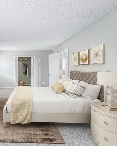 Spacious bedroom with soft gray walls, plush carpeting, and a large upholstered bed dressed in neutral bedding and accent pillows. Matching nightstands with modern lamps frame the bed, while gold-toned wall art adds a contemporary touch. An