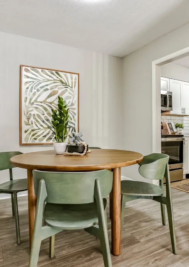 Cozy dining area featuring a round wooden table with green modern chairs, decorative plants, and contemporary wall art. The space includes wood-look flooring, soft neutral walls, and an open view into a bright kitchen with stainless steel a