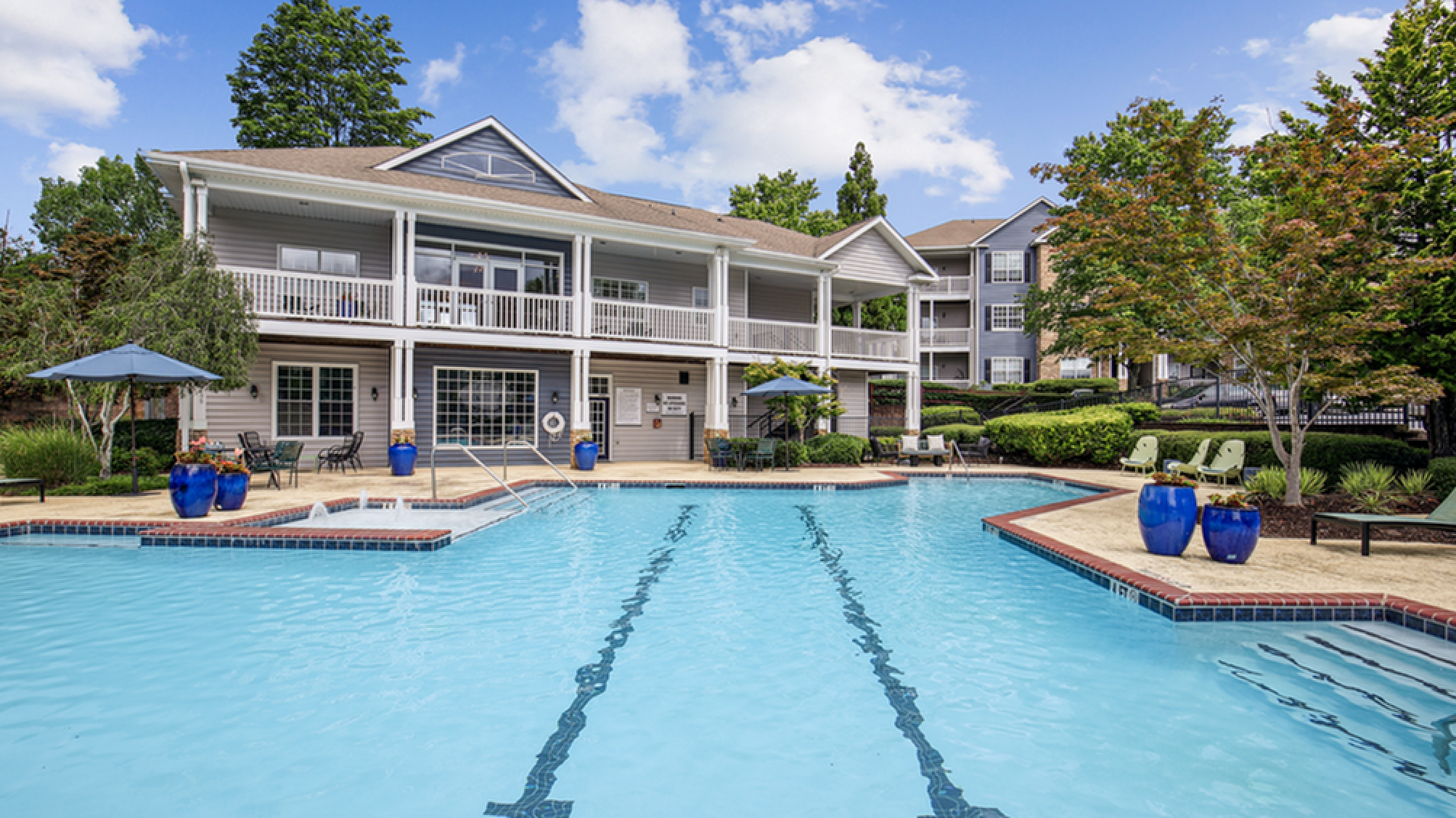 Elegant apartment community with a large, inviting swimming pool, surrounded by lush greenery and stylish outdoor seating.