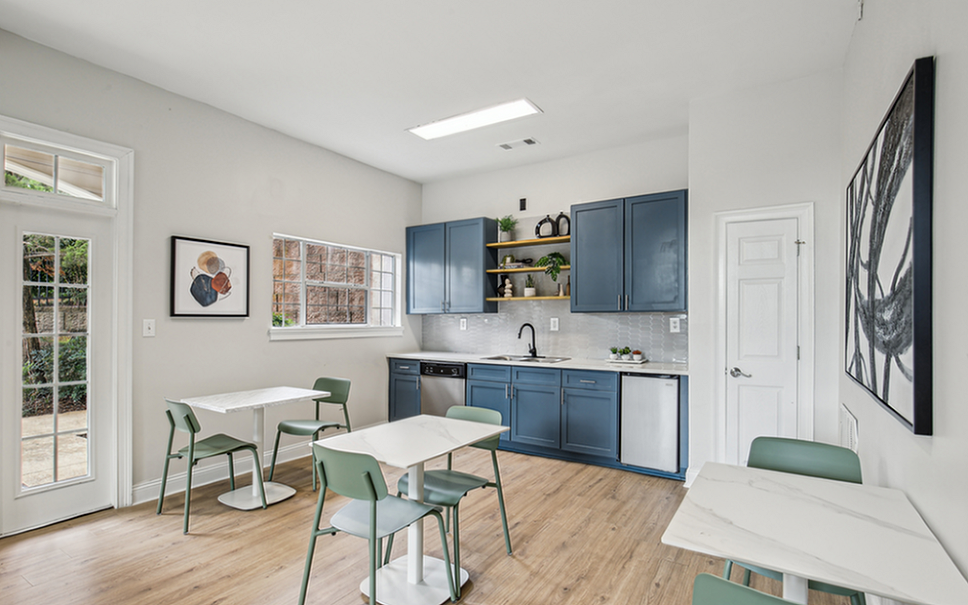 Bright and modern resident area with sleek tables, blue cabinetry, and natural light from large windows.