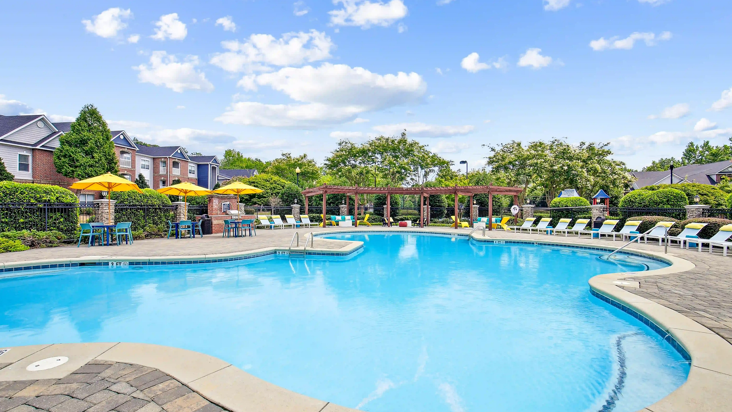Inviting outdoor pool area featuring sun loungers, vibrant yellow umbrellas, and lush greenery under a bright blue sky.