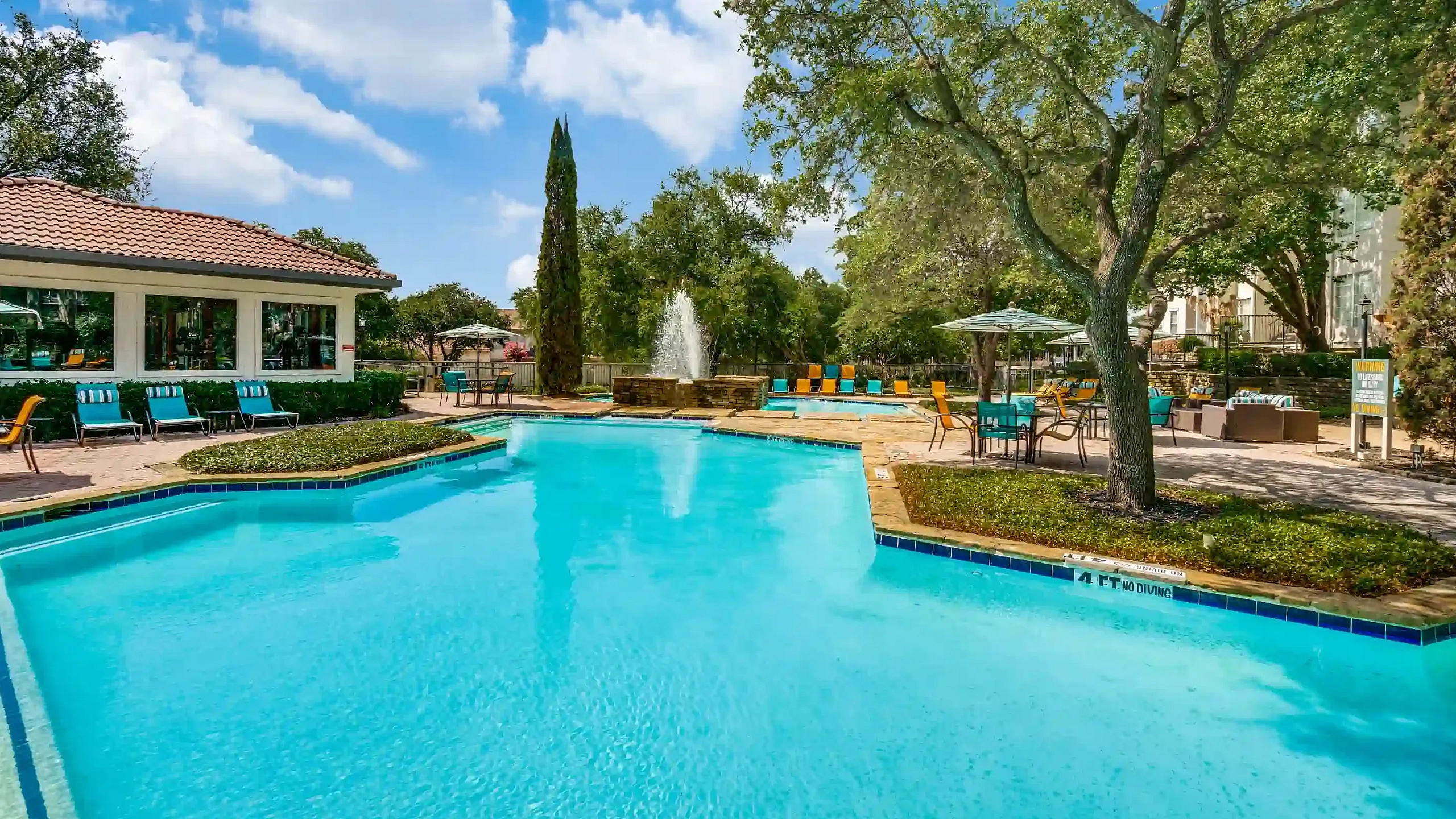 Swimming pool area with sparkling blue water, surrounded by lush greenery, cozy seating, and shaded tables under a sunny sky.
