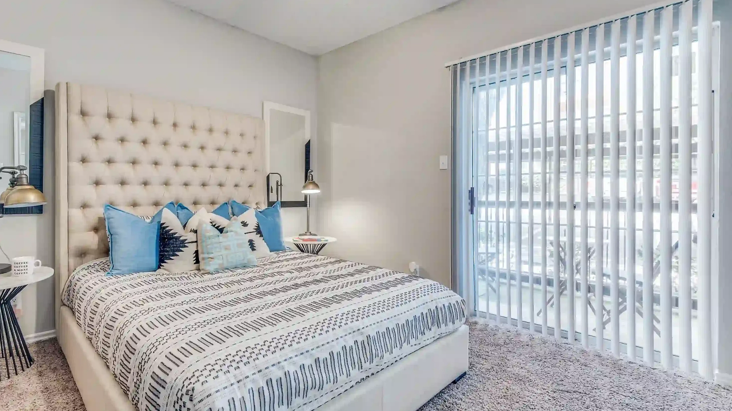 Bright and inviting bedroom with a plush, tufted headboard, stylish decor pillows, and a sliding glass door leading to a cozy outdoor space.