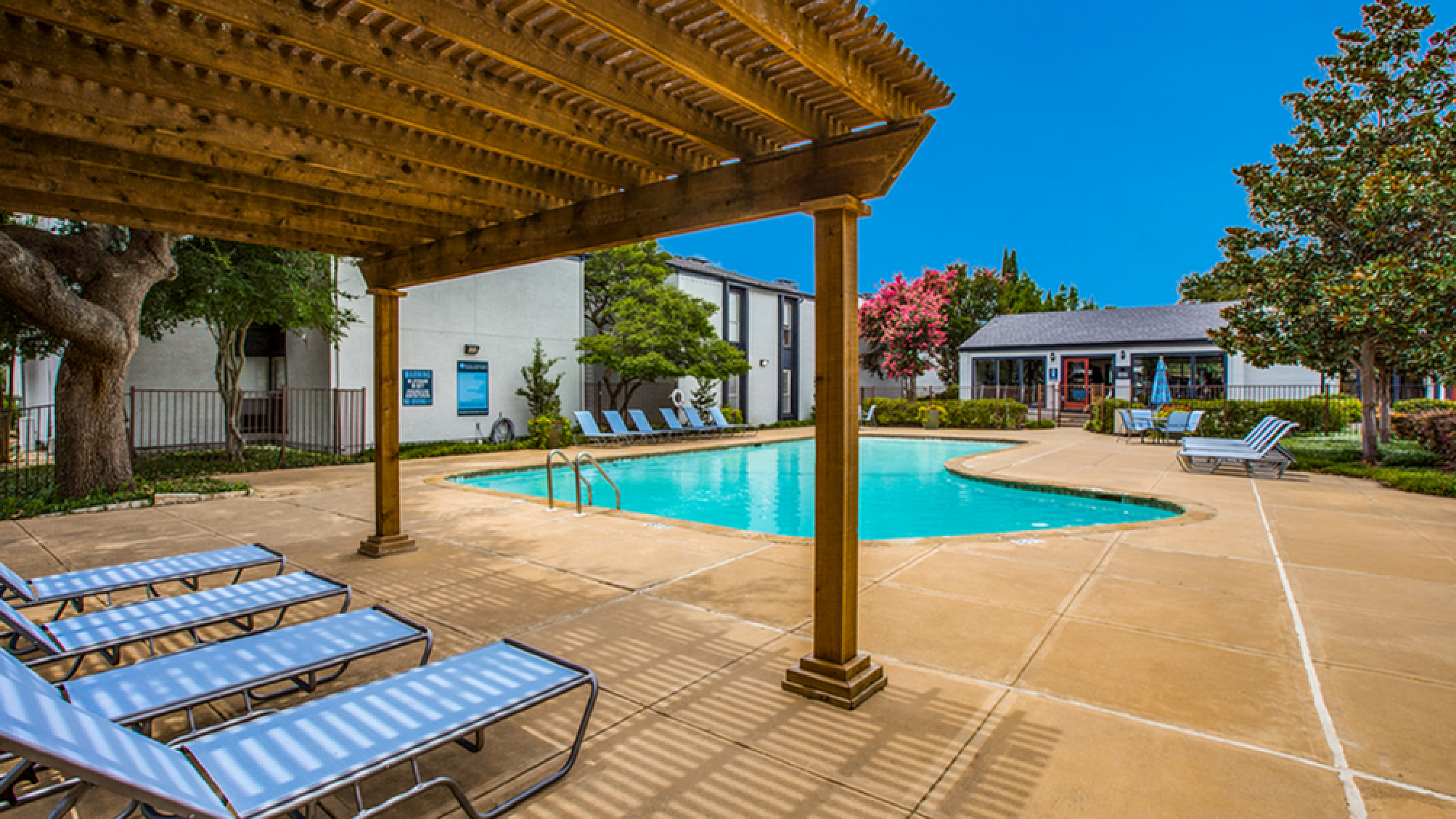 Inviting outdoor pool area with modern loungers and a wooden pergola, surrounded by lush greenery and a beautiful blue sky.