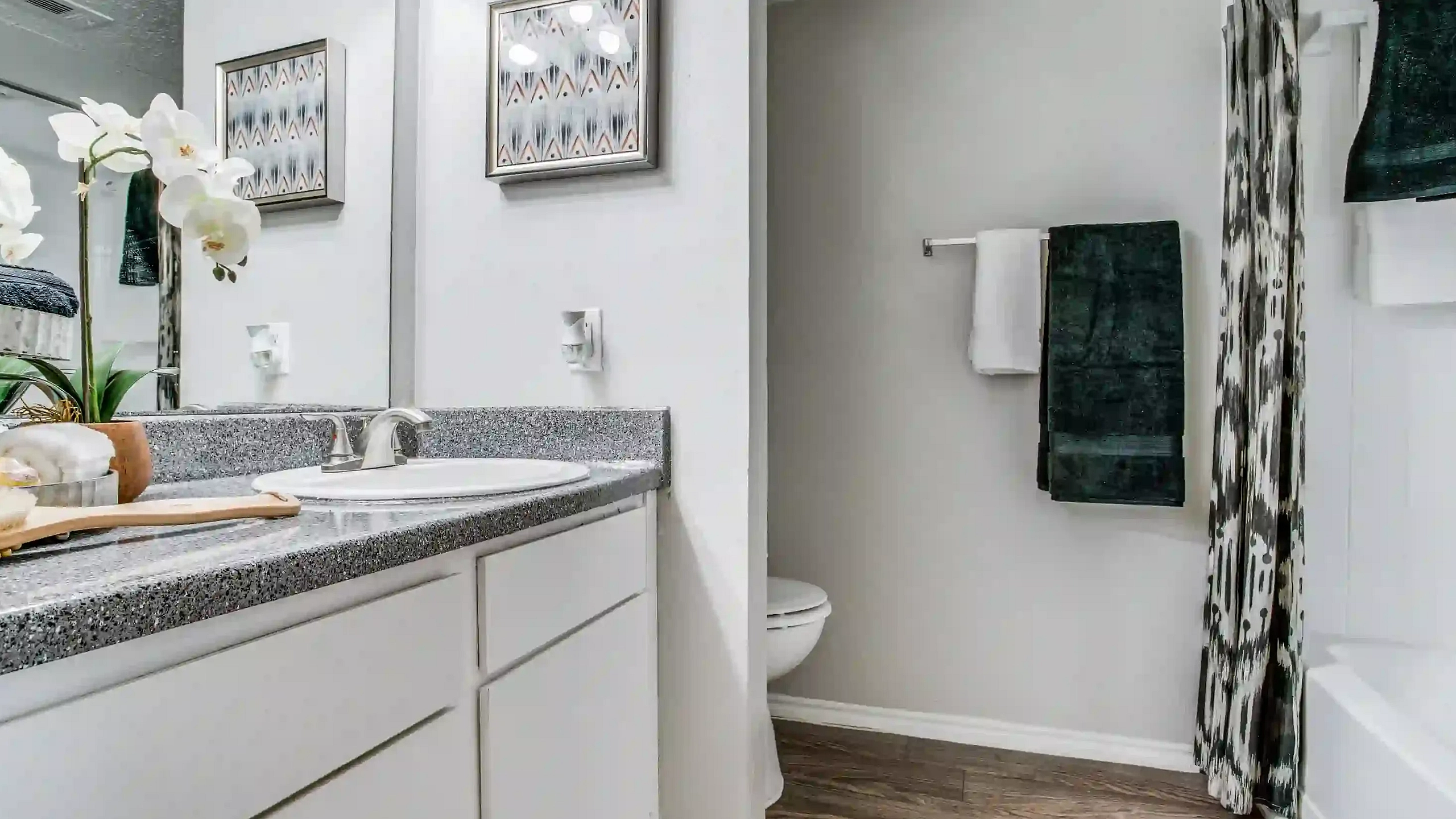 Elegant bathroom with granite style countertop, sleek fixtures, modern artwork, and chic decor featuring lush green towels and a stylish orchid arrangement.