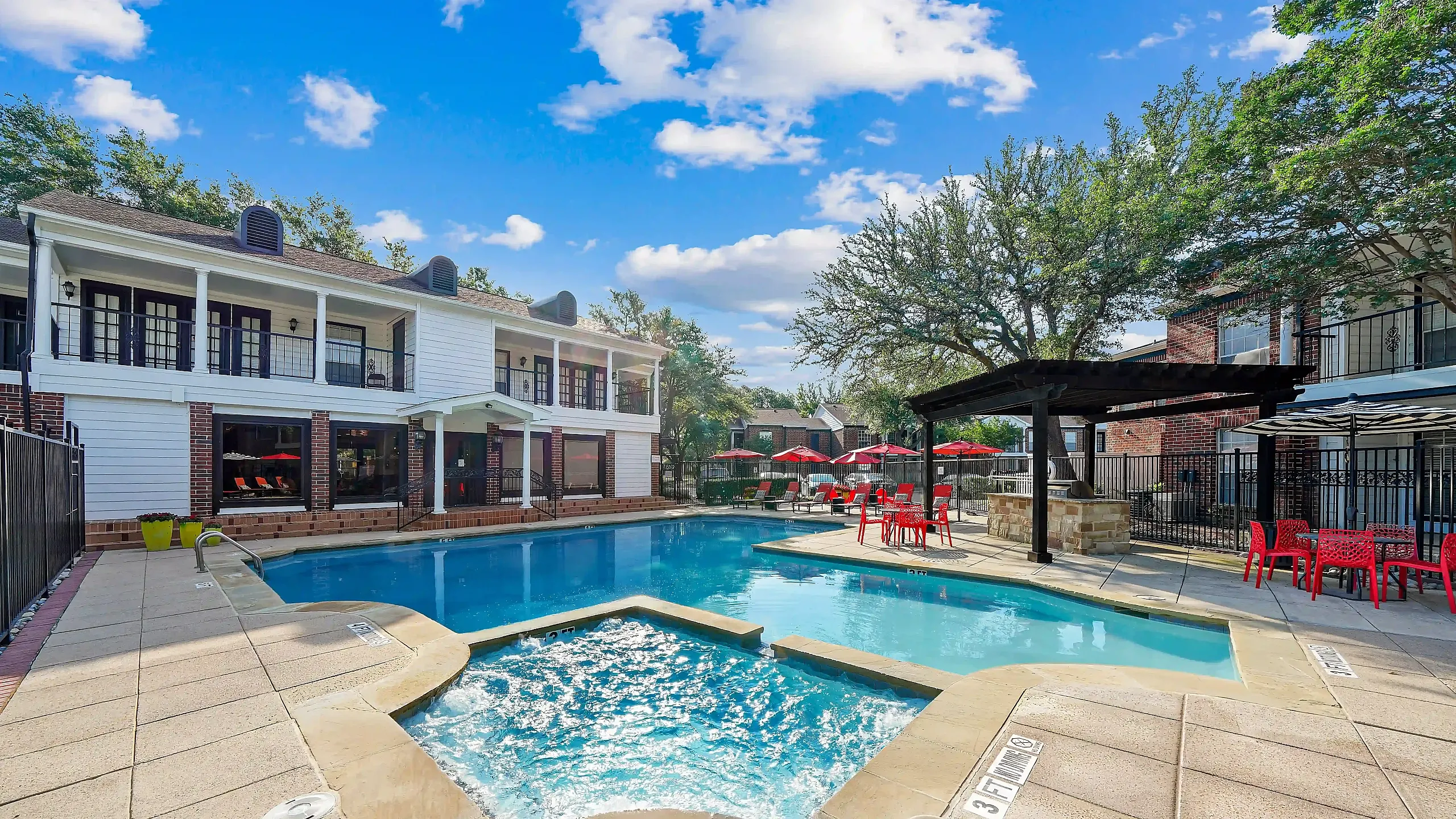 Luxurious apartment community pool area featuring a sparkling swimming pool, relaxing hot tub, and vibrant red seating under the shade of tall trees.