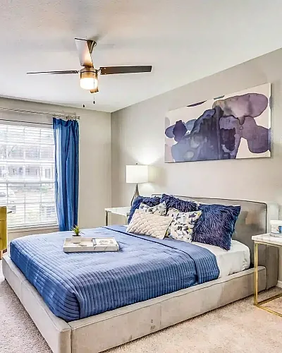 Bright and inviting bedroom with stylish decor, cozy bed with blue accents, and natural light from large window.