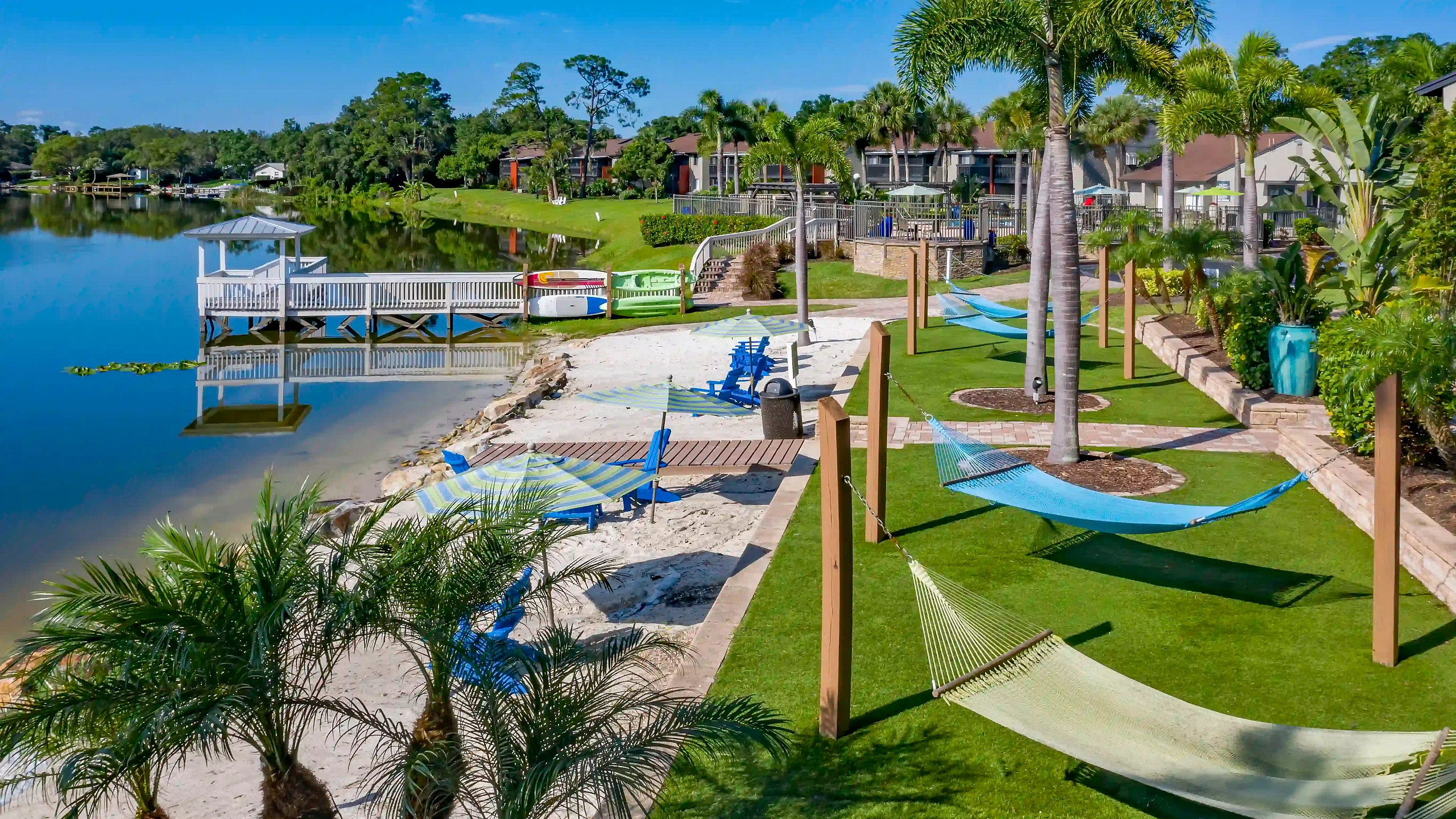 Liv at Winterpark aerial view of a lakeside apartment community featuring a waterfront dock, sandy beach seating area, hammocks, swimming pool, and lush tropical landscaping.