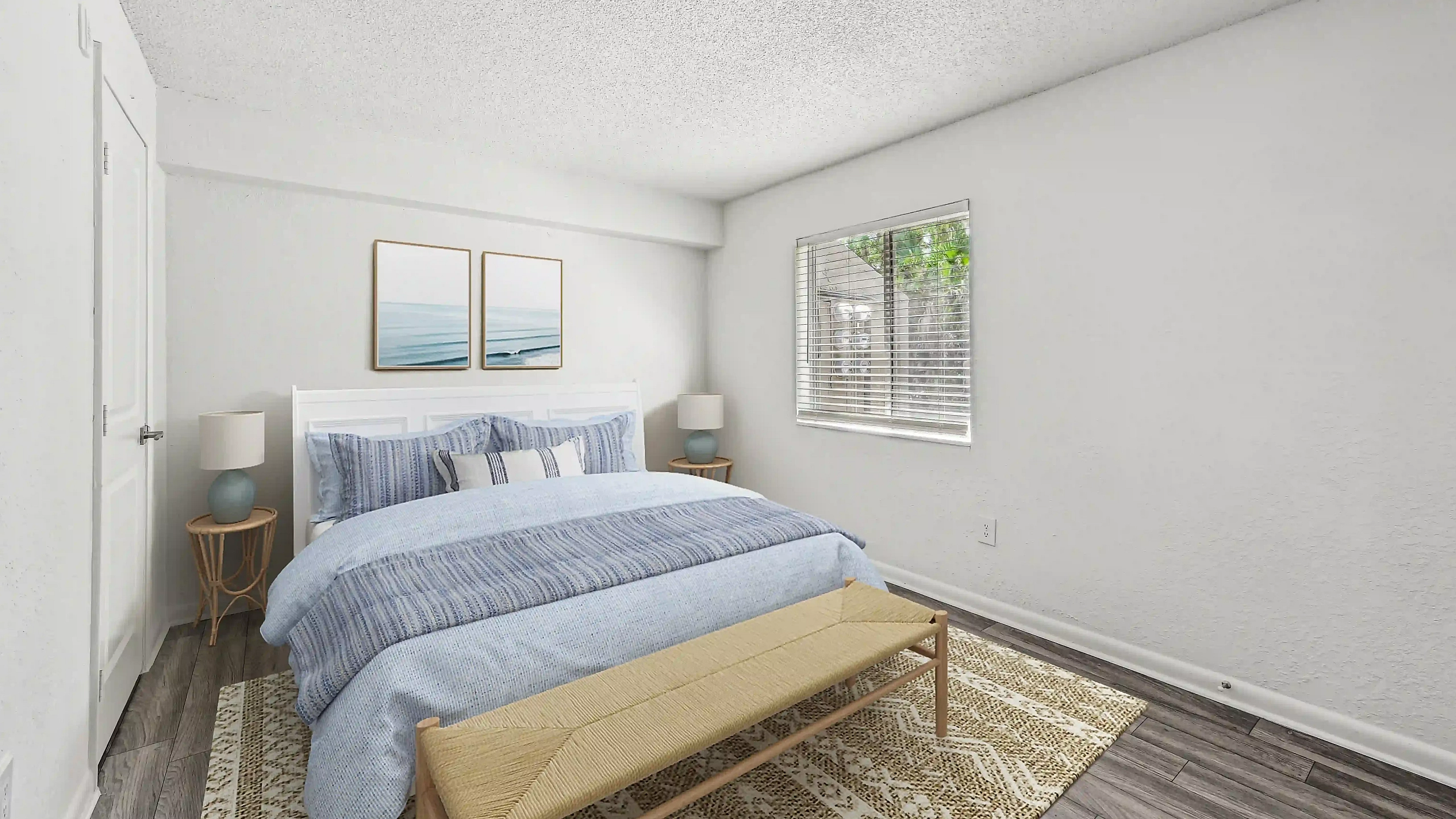 Bright apartment bedroom featuring a bed with blue bedding, matching bedside lamps, a wooden bench, and a window overlooking greenery.