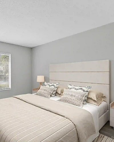 Bright apartment bedroom featuring neutral bedding, a white headboard, matching bedside lamps, a large window with blinds, and wood-look flooring.