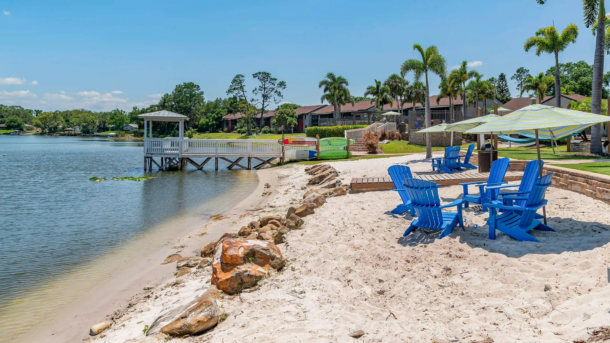 Lakeside apartment community featuring a sandy beach area with blue Adirondack chairs, shaded umbrellas, a waterfront dock, and tropical landscaping.
