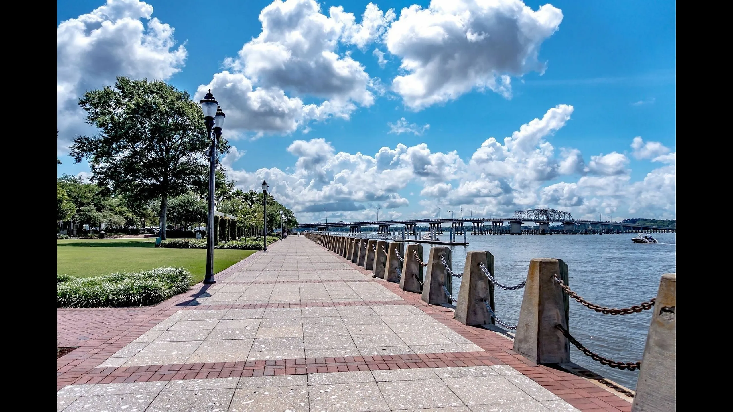 Scenic riverside promenade featuring a lush green park, clear blue skies, and a bridge in the distance, perfect for leisurely walks and enjoying nature.