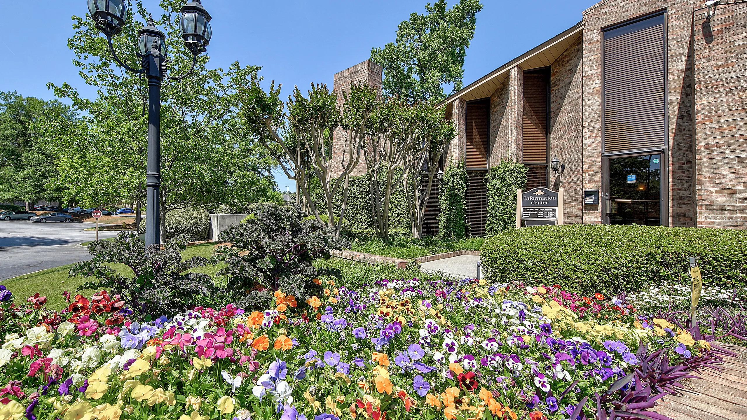 Inviting apartment entrance surrounded by vibrant flowers and lush greenery, featuring a charming brick building with tall windows and a classic street lamp.