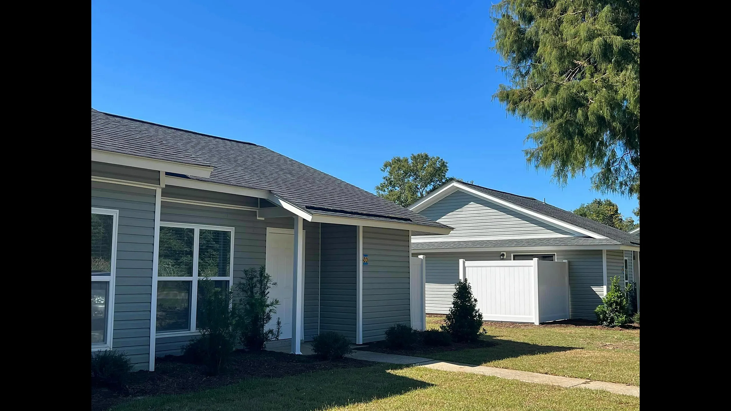 Charming single-story homes with modern gray siding under a clear blue sky, featuring well-maintained lawns and lush greenery.
