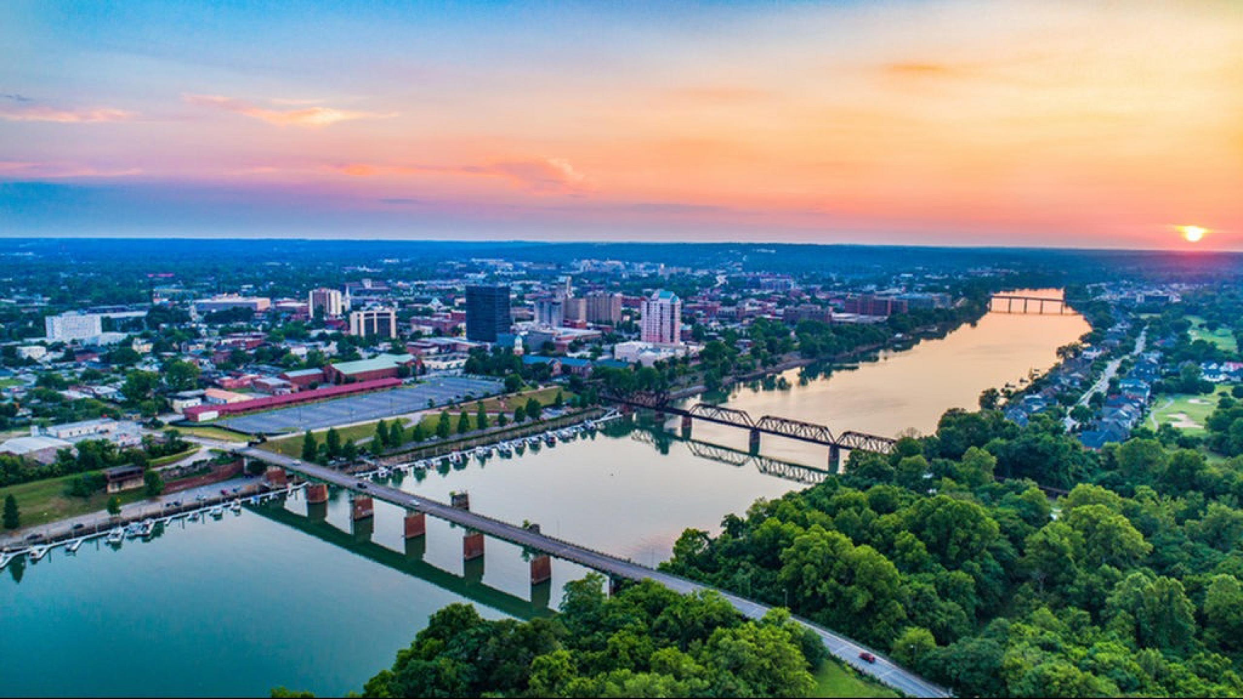 Stunning aerial view of a vibrant city skyline at sunset, featuring a picturesque river with lush green surroundings and a striking bridge.
