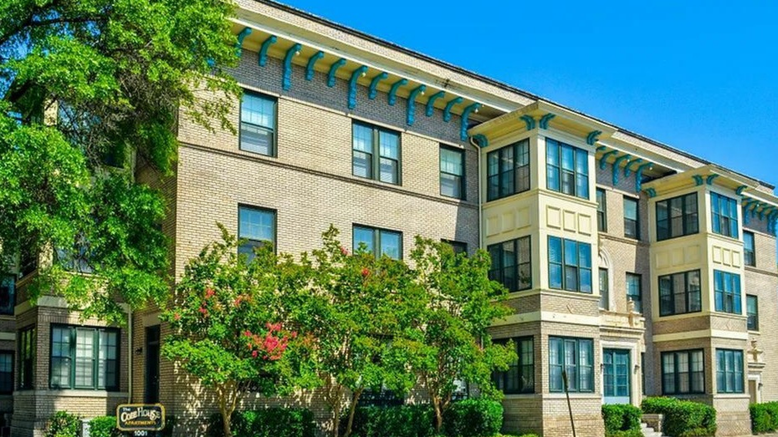 Charming historic-style apartment building with beautiful brick facade, surrounded by lush greenery on a sunny day.