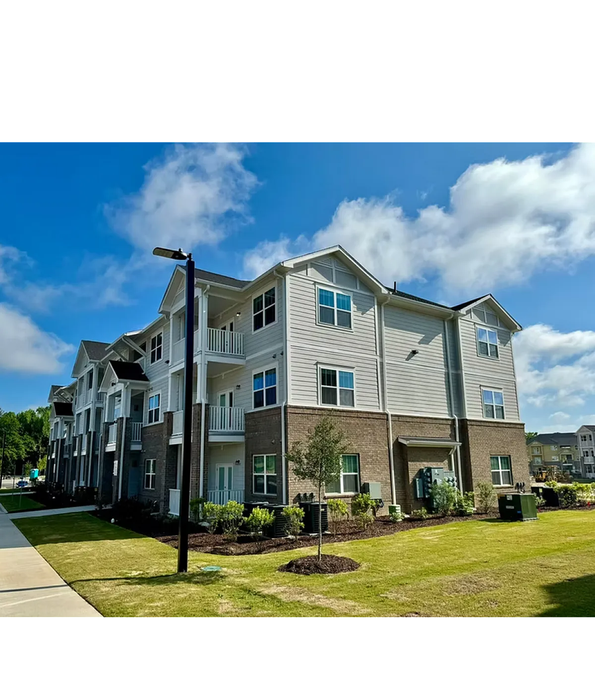 Charming modern apartment building with balconies, nestled in a lush green setting under a bright blue sky.