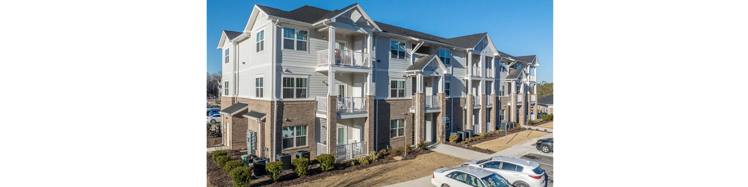 Modern three-story apartment complex with stylish balconies, elegant brick facade, ample parking, and landscaped surroundings under a clear blue sky.