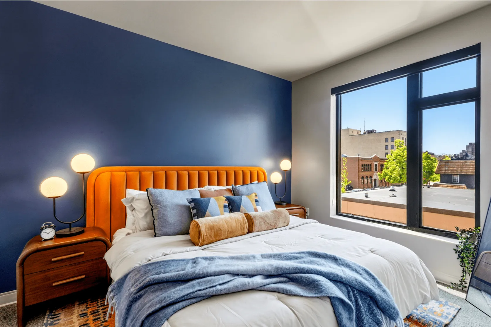 Bright bedroom featuring a plush orange headboard, modern lighting, and a large window with city views.