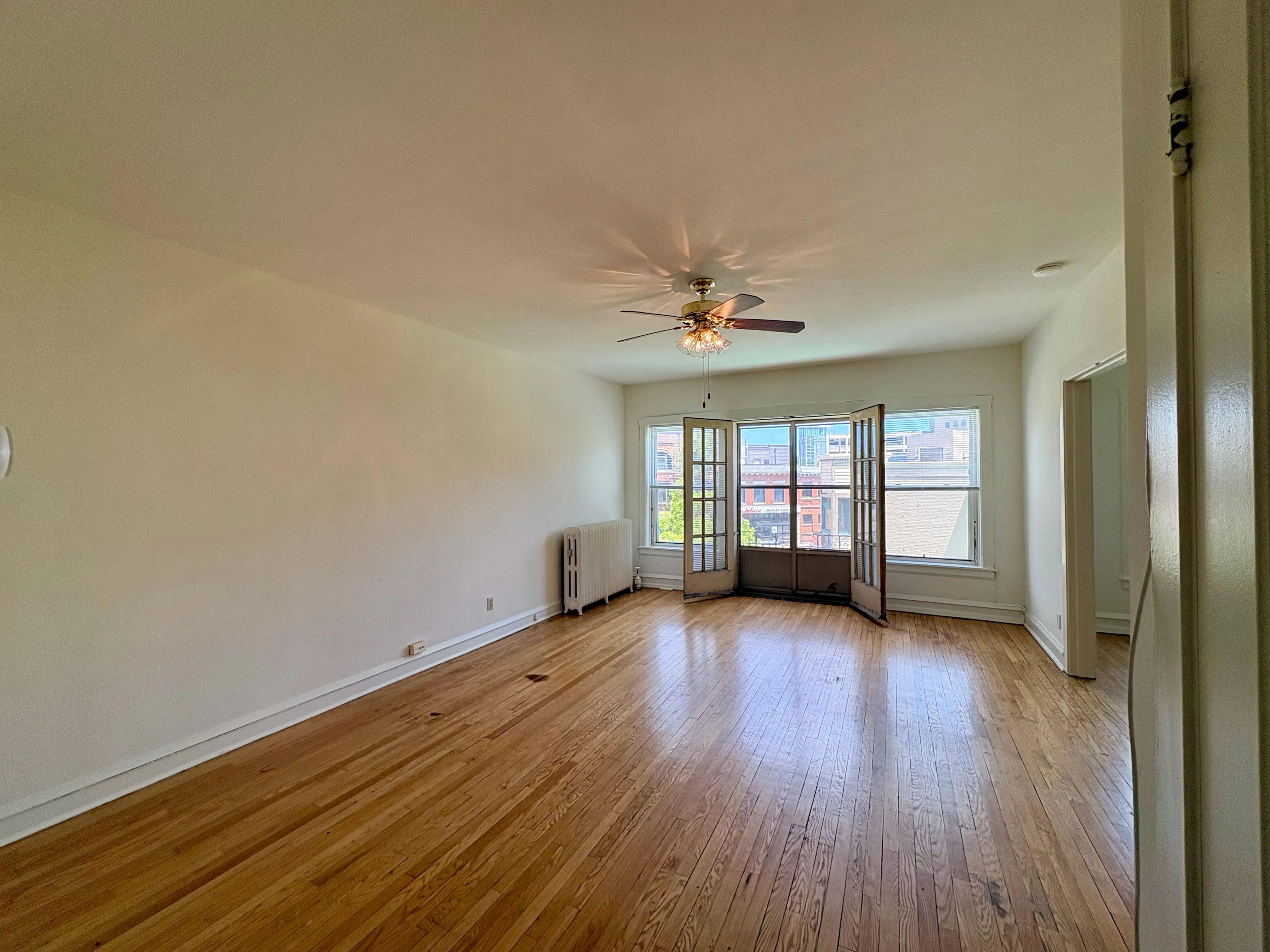 Bright living room with elegant hardwood floors, ceiling fan, and large windows offering city views, perfect for urban living.
