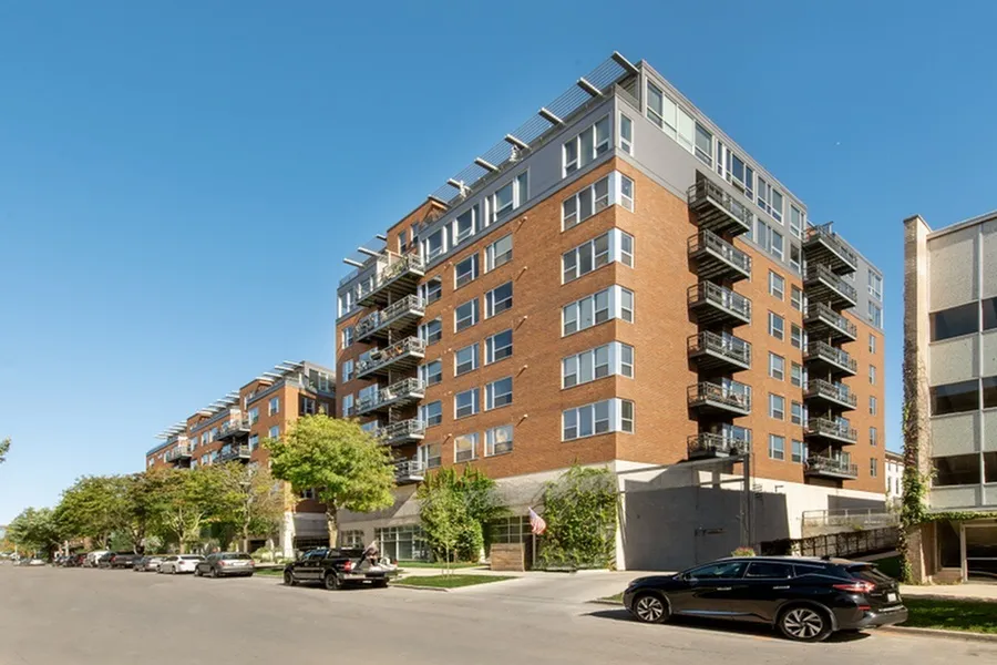 Modern urban apartment building with red brick façade, balconies, and lush greenery in a vibrant city neighborhood.