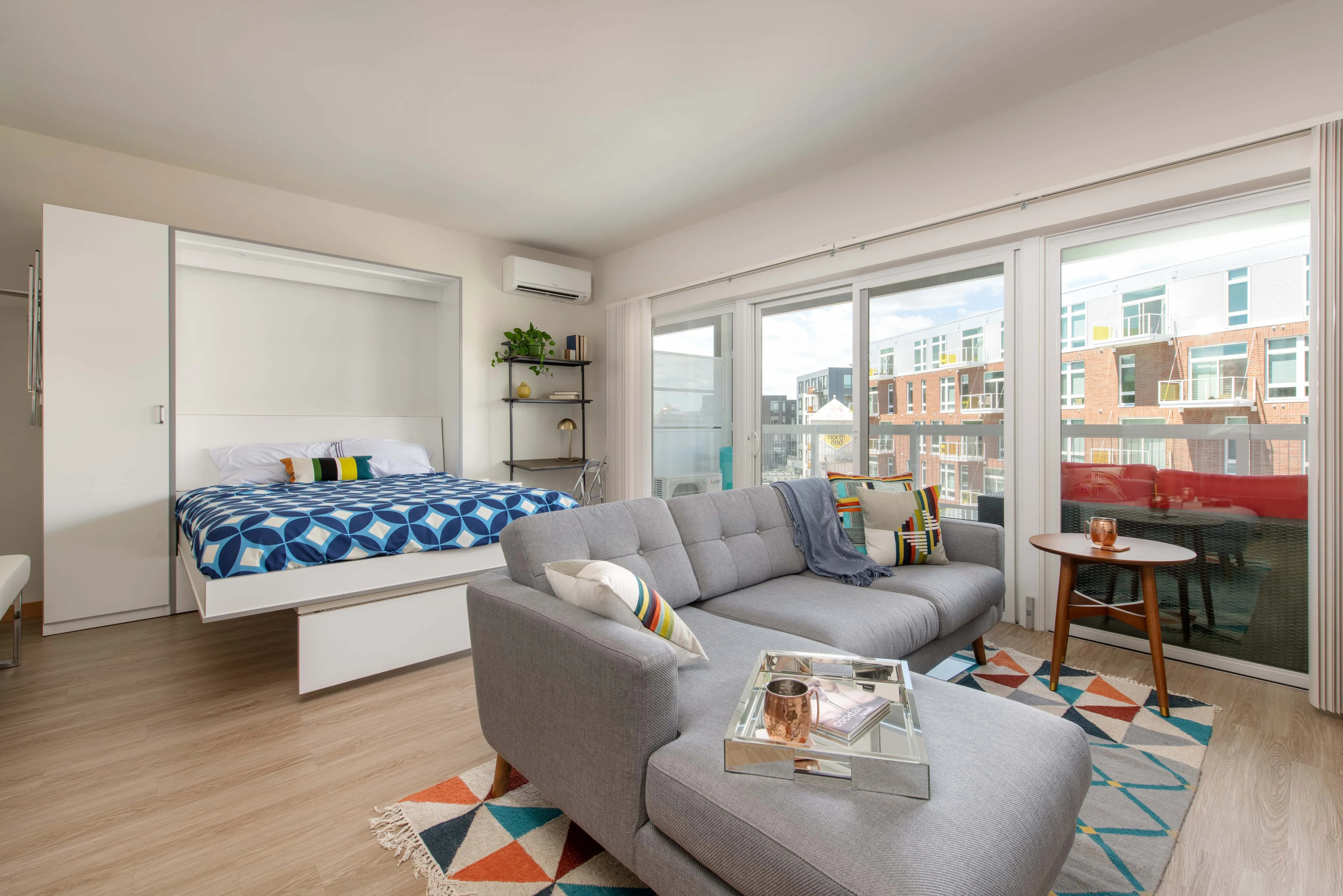Modern studio apartment with a cozy Murphy bed, sleek gray sofa, and vibrant decor, featuring a large balcony with urban views.