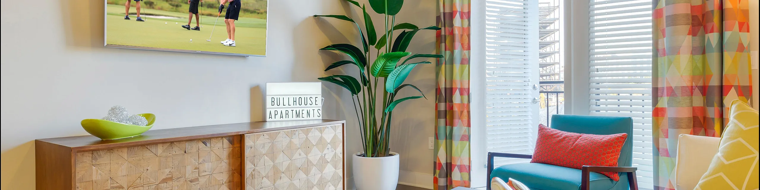 Inviting living room in Bullhouse Apartments featuring modern decor, vibrant geometric curtains, and a cozy seating area with natural light and a lush potted plant.
