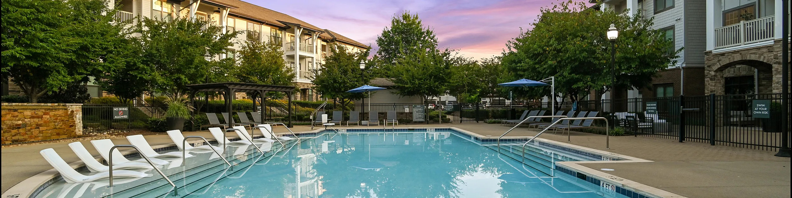 Luxurious apartment complex featuring a stunning swimming pool surrounded by lush greenery, perfect for relaxing evenings under a vibrant sunset sky.