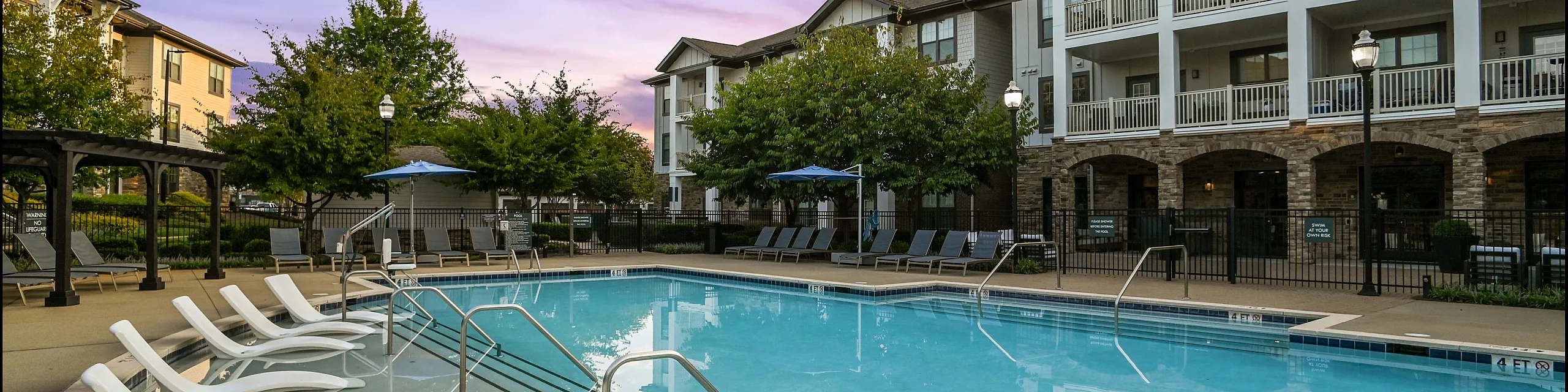 Luxurious apartment complex with a sparkling pool and modern lounge chairs, surrounded by elegant buildings and lush greenery, perfect for relaxing evenings.