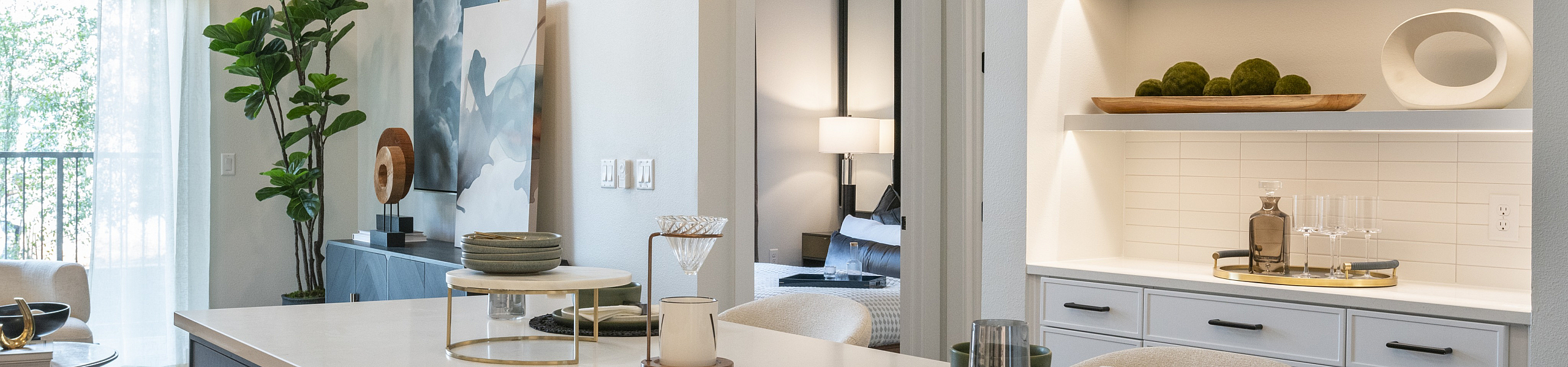 Modern kitchen interior with sleek countertops, stylish shelving, and cozy dining space, leading to a beautifully lit living area with large windows and contemporary decor.