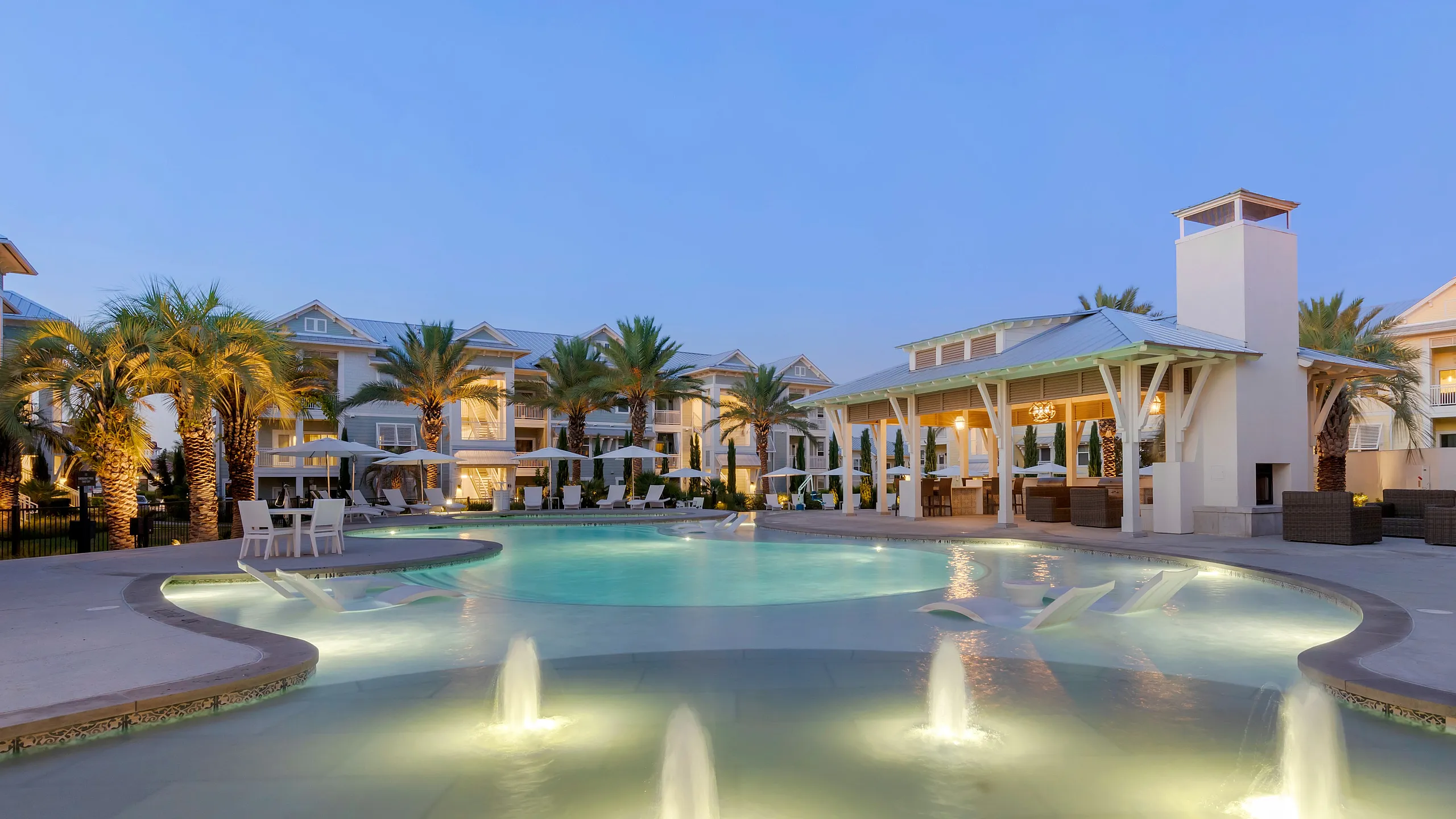 Luxurious resort-style apartment complex featuring a beautifully illuminated swimming pool, elegant lounge chairs, and palm trees under a serene evening sky.