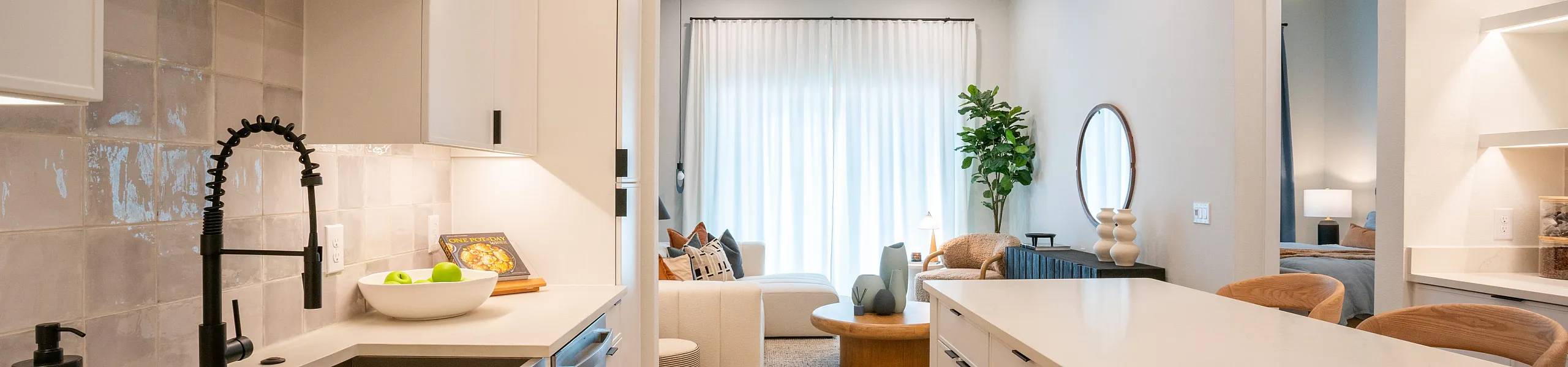 Chic modern kitchen with sleek white cabinetry, spacious island, and open view to a cozy, light-filled living room with elegant decor and lush greenery.