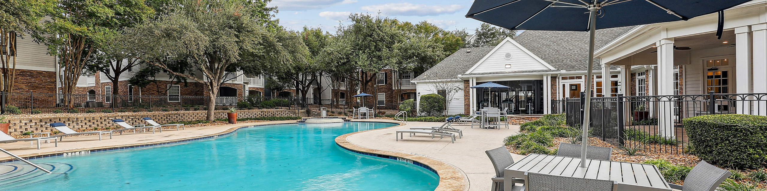 Elegant apartment community pool area with pristine water, sun loungers, and shaded seating under a bright blue sky, surrounded by lush greenery.