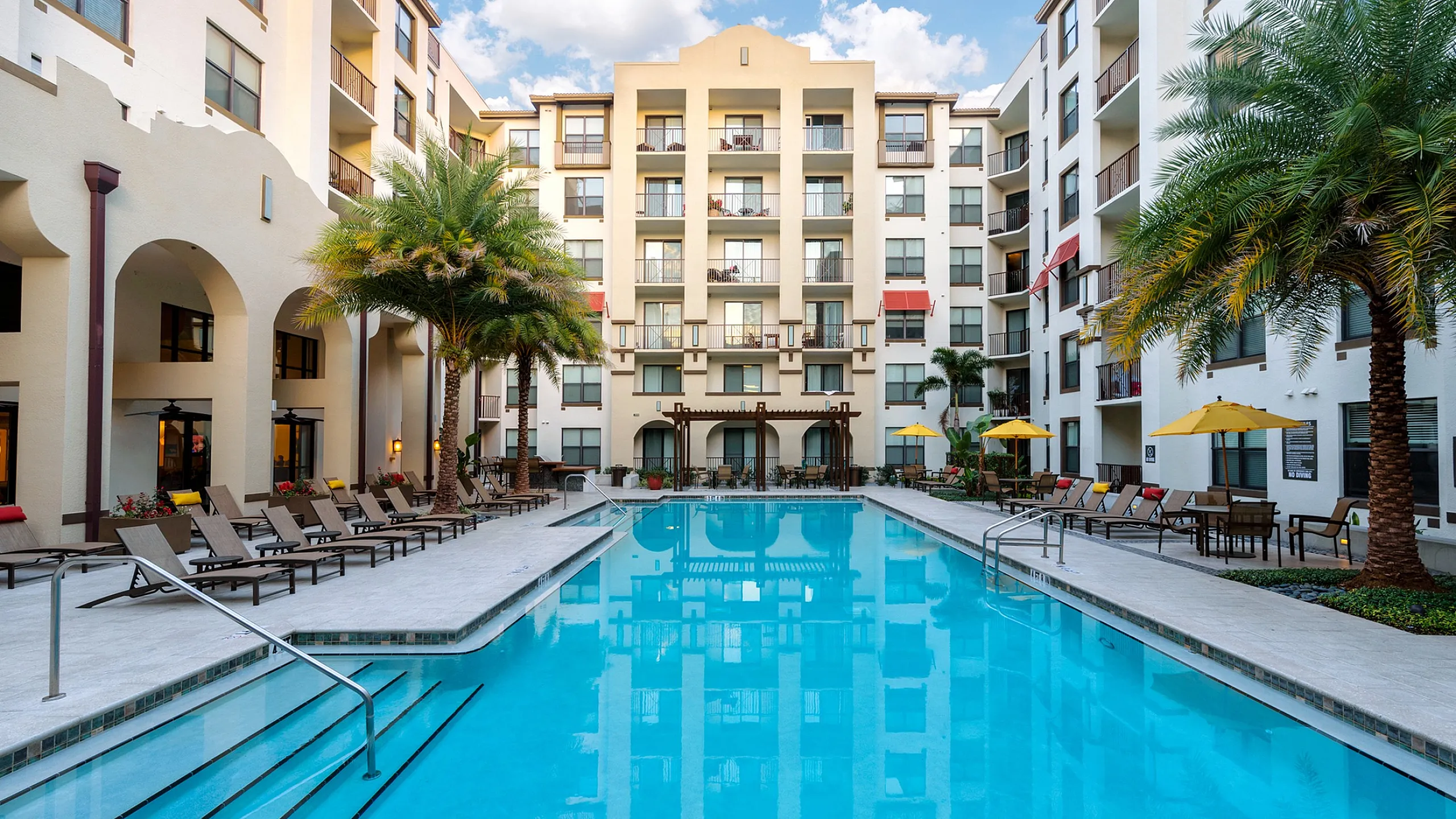 Luxurious apartment complex with a stunning outdoor pool, surrounded by palm trees and elegant lounge chairs, perfect for relaxation.
