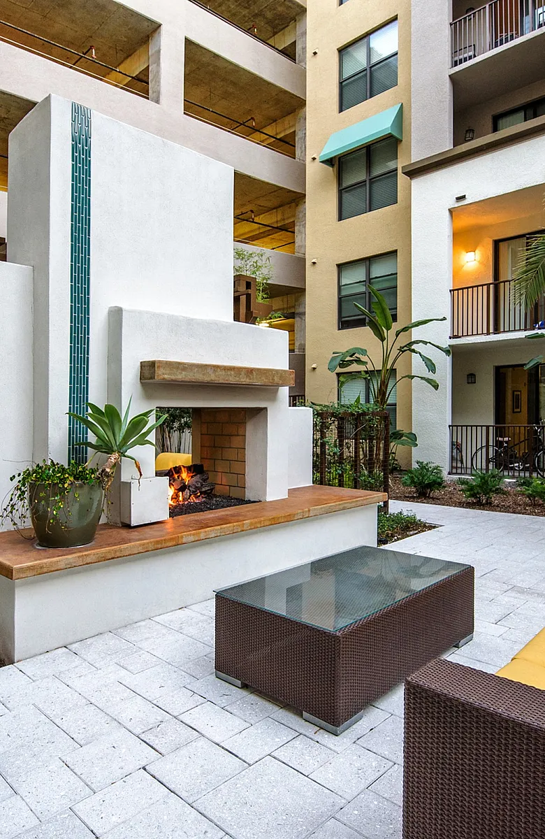 Modern apartment courtyard featuring a cozy outdoor fireplace, stylish wicker seating with bright yellow cushions, and lush greenery, offering a tropical ambiance.