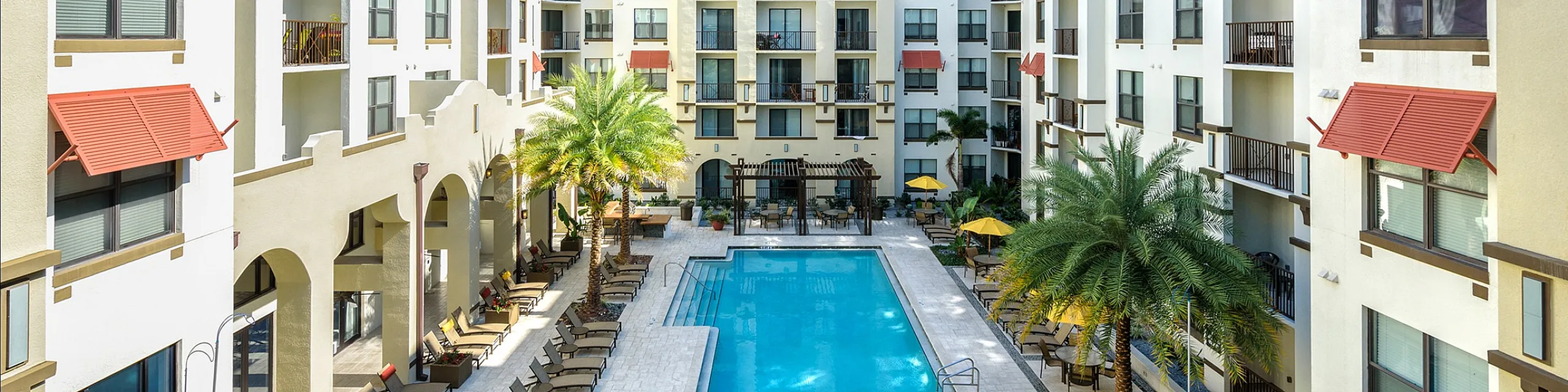 Luxurious apartment complex with elegant architecture, featuring a central sparkling pool surrounded by lush palm trees and relaxing lounge chairs, perfect for a vibrant community lifestyle.