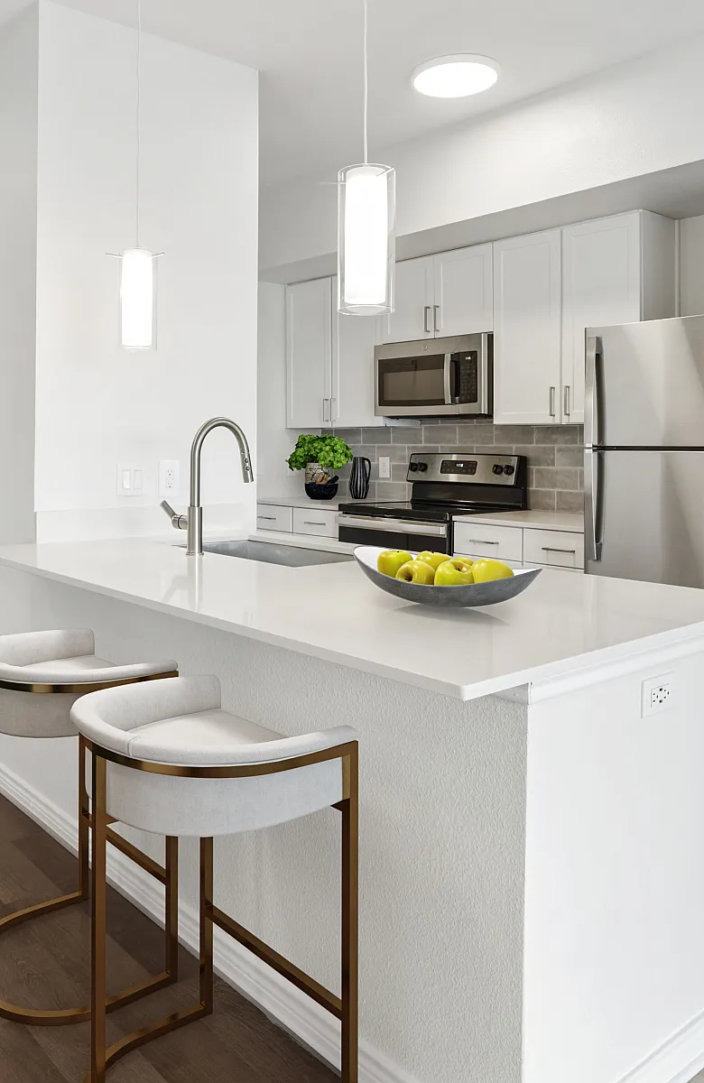 Modern apartment kitchen with sleek white cabinets, stainless steel appliances, and a chic breakfast bar featuring stylish barstools. Adjacent dining area with elegant green chairs and a polished table setting.
