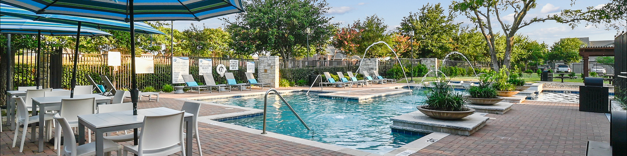Inviting apartment pool area with elegant seating, vibrant umbrellas, and lush greenery under a clear blue sky.