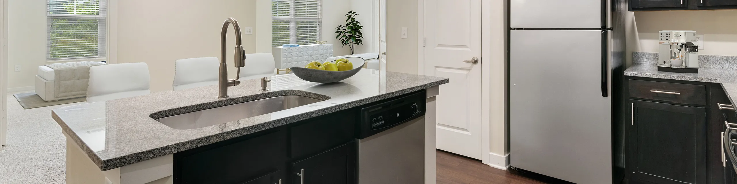 Contemporary kitchen with sleek dark cabinetry, stainless steel appliances, and spacious granite countertops leading to a bright open living area.