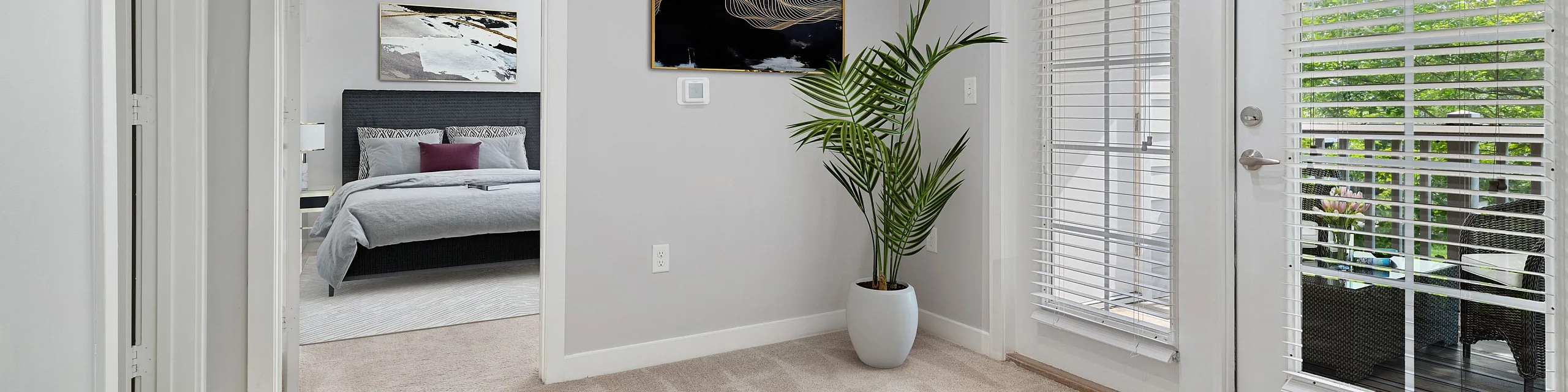 Bright and welcoming apartment foyer with plush carpeting, modern artwork, and access to a serene bedroom and sunlit patio through French doors.