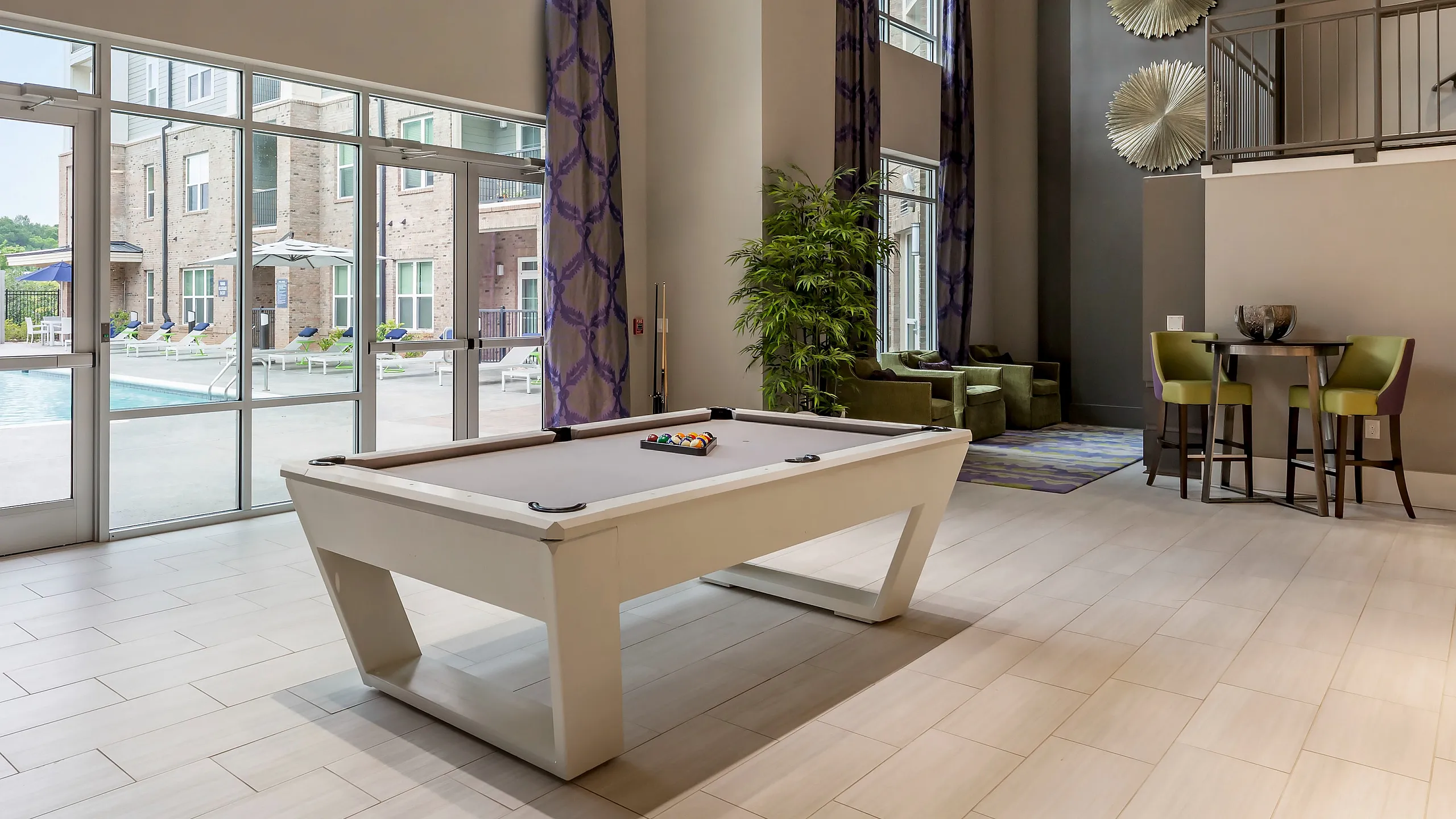 Spacious and modern living area with high ceilings, a stylish pool table, and a view of a luxurious outdoor pool through expansive windows.