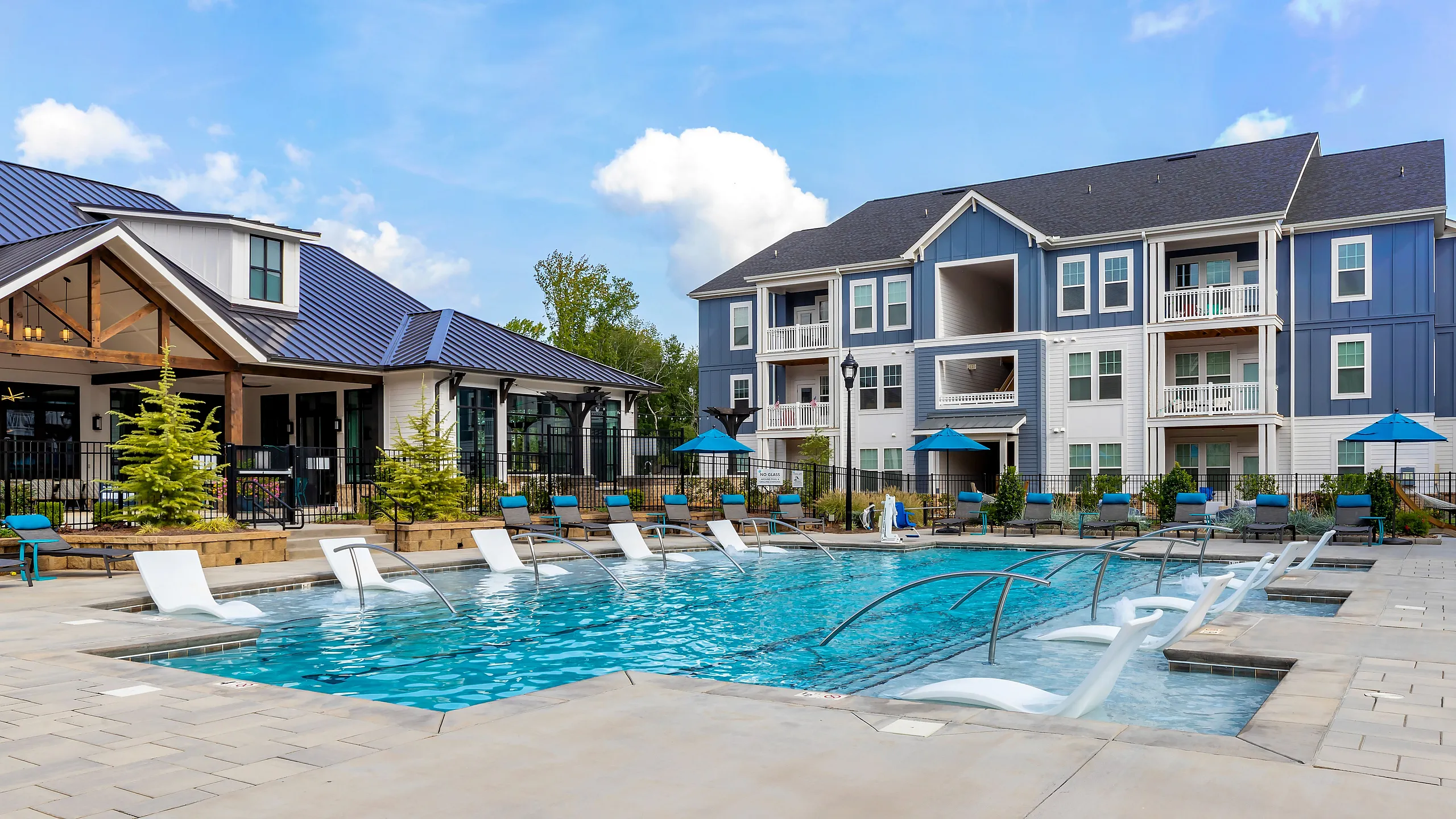 Luxurious apartment complex with a sparkling outdoor pool, stylish lounging chairs, and modern architecture surrounded by lush greenery.