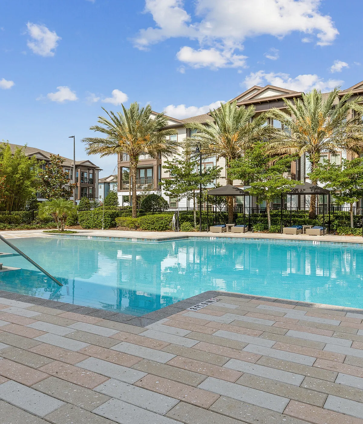 Beautiful apartment complex with a luxurious pool surrounded by tropical landscaping and modern architecture under a clear blue sky.