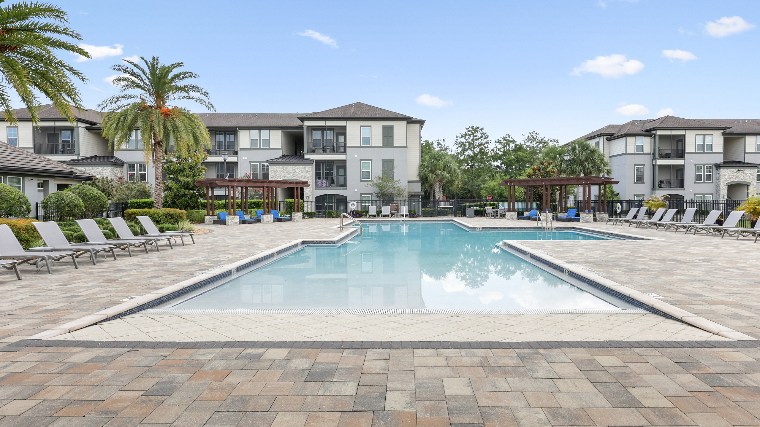 Luxurious apartment complex featuring a stunning swimming pool with lounge chairs, surrounded by palm trees and upscale architecture.