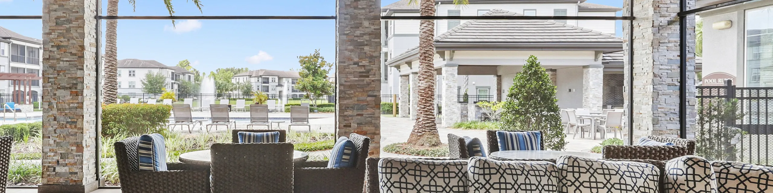 Chic outdoor lounge area with stylish wicker furniture, patterned cushions, and a view of a serene poolside and apartment complex.