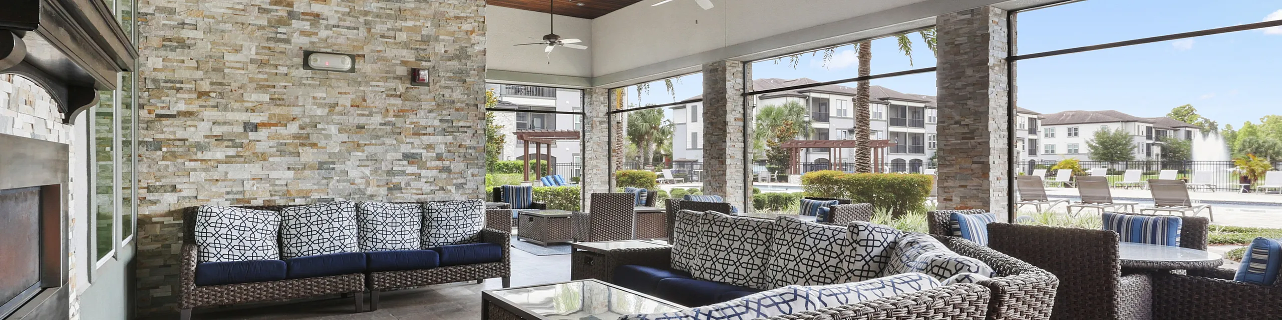Charming outdoor lounge with stylish wicker furniture, stone accents, and ceiling fans, overlooking a tranquil pond and resort-style seating.