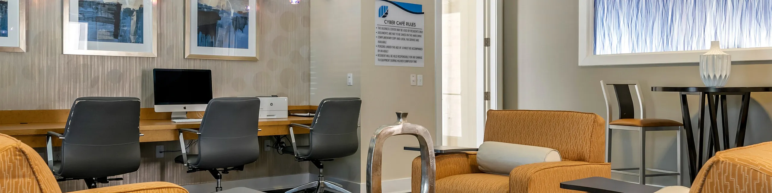 Modern communal lounge area with stylish seating, sleek workstation computers, and vibrant wall art, perfect for relaxation or productivity.