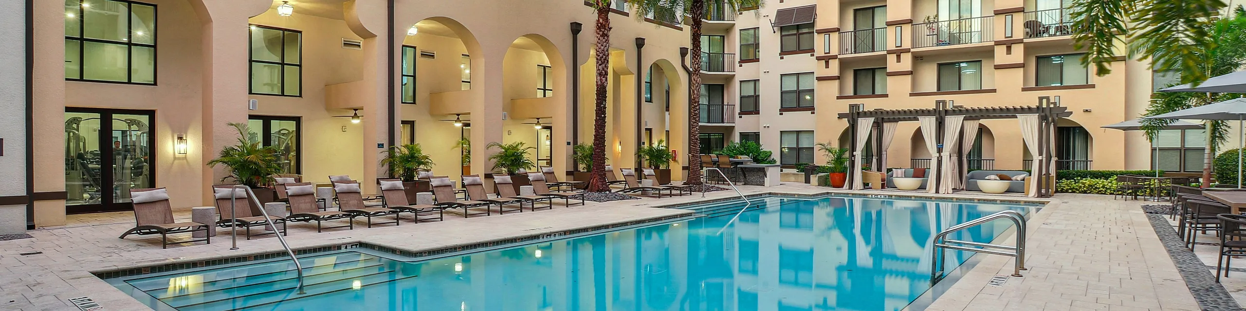 Luxurious apartment complex featuring an inviting outdoor pool surrounded by elegant loungers and swaying palm trees, perfect for relaxation and leisure.