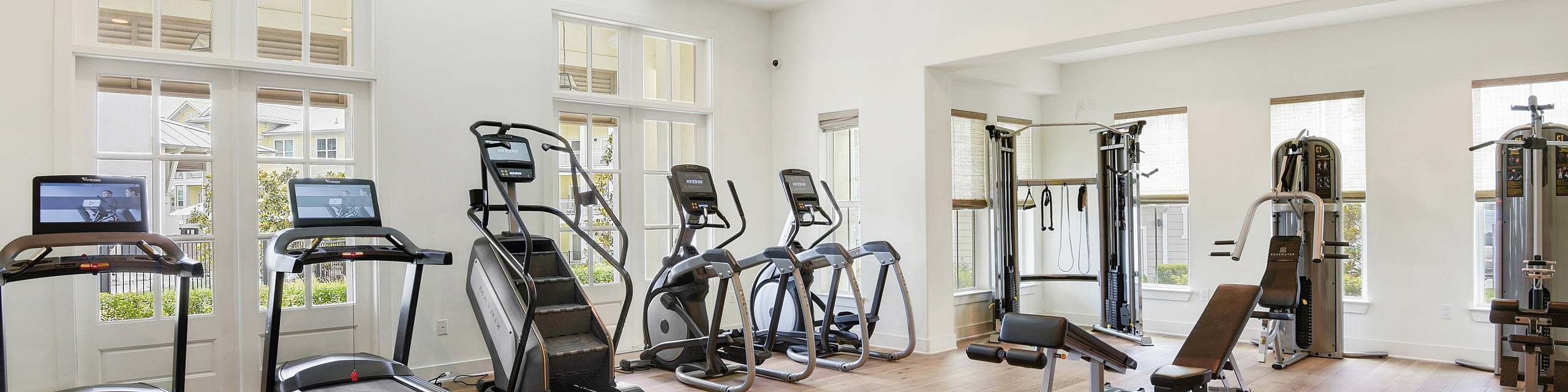 Modern apartment gym with state-of-the-art treadmills, elliptical machines, and strength training equipment, illuminated by natural light through large windows.